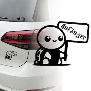 Könnte beinhalten: Eine schwarz-weiße Comic-Schildkröte mit einem breiten Lächeln hält ein Schild mit der Aufschrift "Anfänger" in schwarzer Schrift. Die Schildkröte befindet sich auf einem weißen Auto.