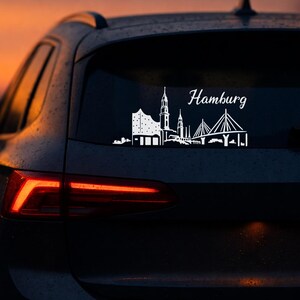Könnte beinhalten: Weißer Vinyl-Aufkleber mit der Skyline von Hamburg, einschließlich Elbphilharmonie, Kirchen und einer Brücke, mit dem Stadtnamen in Schreibschrift. Der Aufkleber befindet sich auf der Heckscheibe eines dunklen Autos vor einem Sonnenuntergang.