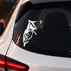 Könnte beinhalten: Ein weißer Vinyl-Aufkleber eines stilisierten Uhus mit scharfen, kantigen Federn und einem fokussierten Blick auf der Heckscheibe eines weißen Fahrzeugs. Das Design zeigt kontrastreiche Linien, die Kopf, Flügel und Körper des Uhus definieren.