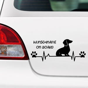 Könnte beinhalten: Ein schwarzer Autoaufkleber aus Vinyl mit der Silhouette eines Dackels, der auf einer Herzschlaglinie sitzt. Das Design enthält Pfotenabdrücke an beiden Enden der Linie sowie den Text WUNSCHNAME on BOARD über der Herzschlag-Grafik.