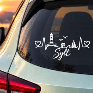 Könnte beinhalten: Ein weißer Vinyl-Aufkleber auf einer Autoscheibe mit einem durchgehenden Herzschlag-Design. Die Grafik zeigt einen Leuchtturm, einen Strandkorb, ein Segelboot, zwei Vögel und zwei Herzen, mit dem Wort Sylt in einer geschwungenen Schreibschrift.