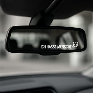 Puede incluir: Espejo retrovisor negro con el texto blanco "ICH HASSE MENSCHEN" y un gráfico de cara de diablo. El espejo está dentro de un coche, con el techo y el salpicadero visibles.