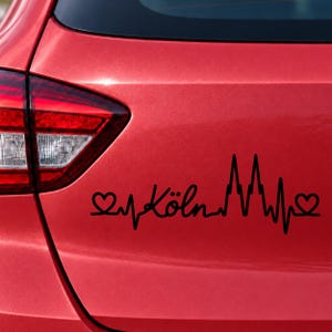 Könnte beinhalten: Ein schwarzer Vinyl-Aufkleber auf dem Kofferraum eines roten Autos. Das Design zeigt eine durchgehende Herzschlaglinie mit einem Herzsymbol an beiden Enden, dem Wort Köln in Schreibschrift und der Silhouette der Kölner Domtürme.
