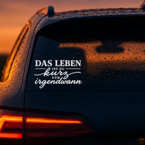 Könnte beinhalten: Ein weißer Vinyl-Aufkleber auf einer Heckscheibe mit dem deutschen Text "DAS LEBEN IST ZU kurz FÜR irgendwann", der in einer Kombination aus fetter Blockschrift und einer fließenden Schreibschrift auf der Glasfläche zentriert ist.
