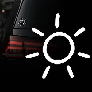 Könnte beinhalten: Weißer sonnenförmiger Aufkleber auf einer schwarzen Autoscheibe. Das Design umfasst einen zentralen Kreis mit Strahlenlinien, und ein kleineres Sonnendesign ist ebenfalls sichtbar. Das Bild ist eine Nahaufnahme, die den Kontrast hervorhebt.