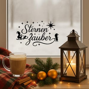 Può includere: Una scena natalizia accogliente con una lanterna di metallo scuro con una candela accesa, una tazza di caffè, bastoncini di cannella e decorazioni dorate. La finestra mostra il testo "Sternenzauber" con illustrazioni di stelle ed elfi.