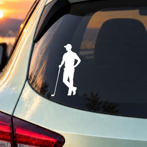 Könnte beinhalten: Ein weißer Vinyl-Aufkleber auf einer Autoscheibe, der die Silhouette eines Golfspielers mit Kappe und Poloshirt zeigt, der mit einer Hand in der Hüfte steht und einen Golfschläger hält, der neben ihm auf dem Boden ruht.