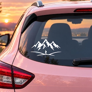 Könnte beinhalten: Ein weißer Vinyl-Aufkleber auf der Heckscheibe eines rosafarbenen Autos. Der Aufkleber zeigt eine grafische Illustration eines Gebirges mit der Silhouette einer wandernden Person auf einem Pfad im Vordergrund unter den Gipfeln.