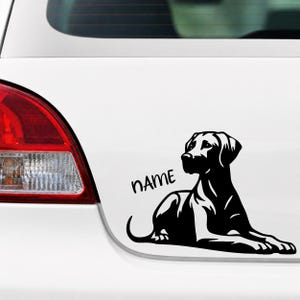 Könnte beinhalten: Ein schwarzer Vinyl-Aufkleber eines liegenden Hundes in Ruheposition, mit dem Wort NAME in einer stilisierten schwarzen Schrift über dem Rücken des Hundes. Der Aufkleber ist auf der weißen Oberfläche eines Fahrzeugs nahe dem Rücklicht angebracht.