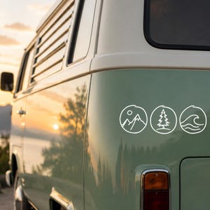 Könnte beinhalten: Ein weißer Vinyl-Aufkleber mit drei kreisförmigen Strichzeichnungen an der Seite eines hellgrünen Vans. Die Symbole zeigen ein Gebirge mit Sonne, einen Nadelbaum und Meereswellen, alles in einem einfachen, klaren weißen Umriss-Stil gehalten.