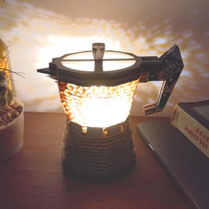 Op de afbeelding: Een decoratieve lamp in de vorm van een koffiepot, gemaakt van karton. De lamp straalt een warme, uitnodigende gloed uit. De handgreep heeft een patroon. De lamp staat op een houten oppervlak.