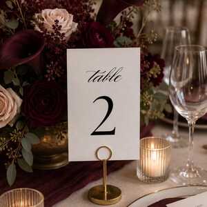 May include: A table setting with a floral arrangement and a table number card. The card reads "table 2" in elegant script. The flowers are burgundy and blush roses and calla lilies. A gold candle holder and wine glasses are also present.