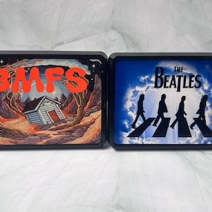 May include: Two black rectangular light-up signs. One features the letters "BMFS" in orange over a landscape with a house. The other sign displays "The Beatles" and silhouettes of the band members crossing a street.