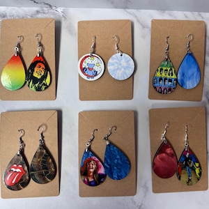 May include: Six pairs of dangle earrings with various designs. The earrings are displayed on brown card stock. The designs include a tie-dye pattern, a cartoon bear, a red and green ombre, a camouflage pattern, a red tongue, a group of people, and a blue and white marble pattern.
