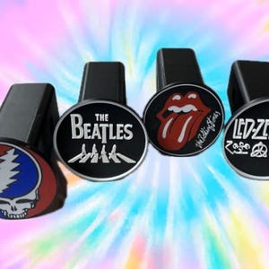 May include: Four black metal trailer hitch covers with circular faces featuring iconic rock band logos. The designs include the Grateful Dead skull, The Beatles text, The Rolling Stones tongue and lips, and the Led Zeppelin symbols and text.