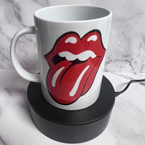 Puede incluir: Taza de cerámica blanca con un logotipo de labios rojos y negros. La taza tiene un asa curva y se muestra sobre un soporte giratorio negro. El diseño de los labios es una imagen audaz e icónica.