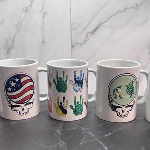 May include: Five white ceramic mugs with different designs. The mugs feature a skull with an American flag, a skull with a green and white design, a skull with a green and white design, a skull with a green and white design, and a building with the text "Train Station".
