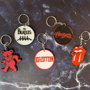 May include: A collection of keychains featuring iconic band logos. Keychains include The Beatles, Aerosmith, Led Zeppelin, and the Rolling Stones' tongue logo. Each keychain has a silver ring and chain.