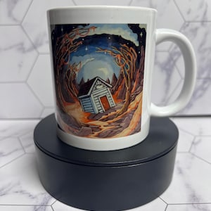 May include: White ceramic mug featuring a detailed illustration of a small house in a surreal landscape. The artwork includes a night sky, trees, and a path leading to the house. The mug has a curved handle and is set on a black stand.