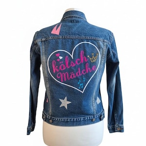 Könnte beinhalten: Eine blaue Jeansjacke mit einer großen weißen Herzumrandung auf der Rückseite. Im Herz steht der Text "kölsch Mädche" in Pink, ergänzt durch eine goldene Krone, kleine Herzen und blaue Musiknoten. Silberne Sterne verzieren den unteren Rückenbereich.