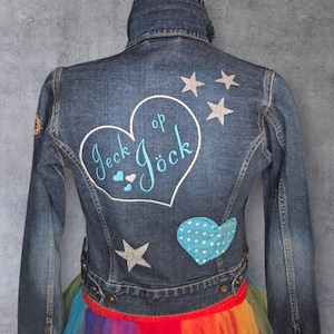 Könnte beinhalten: Eine Jeansjacke mit herzförmigem Design auf dem Rücken, mit dem Text "Jeck op Jöck" in türkisblauer Schrift. Die Jacke ist mit silbernen Glitzersternen und einem blau gepunkteten Herzen verziert. Am unteren Rand ist ein Regenbogenrock zu sehen.