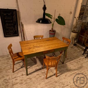 Könnte beinhalten: Ein rustikaler Esstisch aus Holz mit grün lackiertem Untergestell und einer Schublade. Vier Holzstühle umgeben den Tisch. Eine schwarze Industrie-Leuchte hängt darüber. Der Raum hat eine Vintage-Ästhetik.