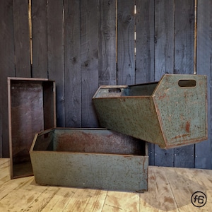 Könnte beinhalten: Drei Vintage-Metall-Aufbewahrungsboxen in verschiedenen Größen. Die Boxen sind in einem verwitterten Teal-Farbton lackiert, mit sichtbaren Rostflecken, die auf Alter und Nutzung hindeuten. Jede Box hat eine rechteckige oder trapezförmige Form mit ausgeschnittenen Griffen an den Seiten.