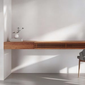 Peut inclure: Un bureau moderne en bois avec un tiroir int&eacute;gr&eacute; et un design &agrave; lattes. Un petit vase blanc avec des tiges noires et une pile de livres sont pos&eacute;s sur le bureau. Une chaise grise avec des pieds en bois est &agrave; proximit&eacute;. Le bureau est contre un mur blanc.