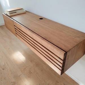 May include: A light wood floating shelf with a slatted front and a small hole on the top surface. The shelf is mounted on a white wall and has a few books on top.