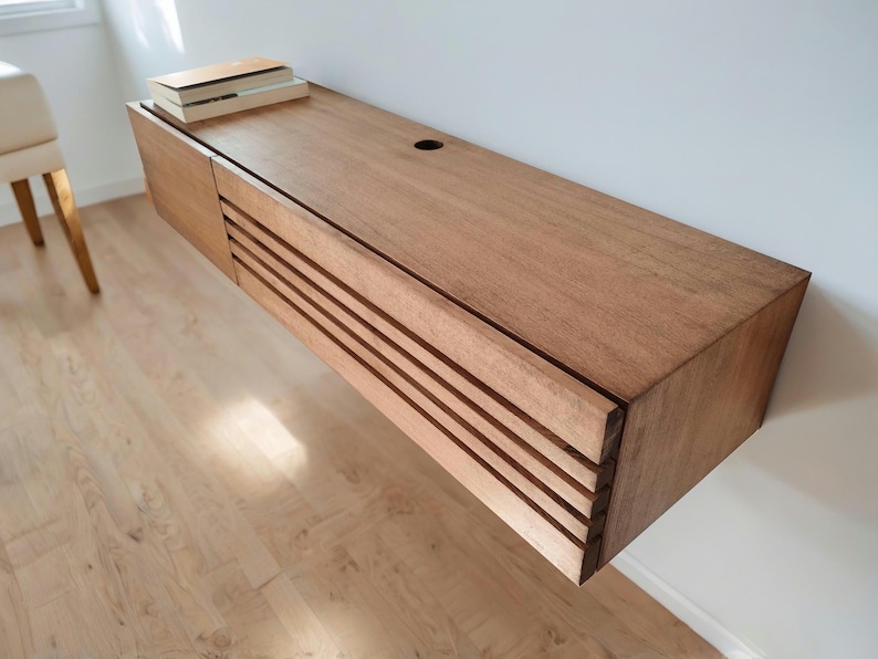 May include: A light wood floating shelf with a slatted front and a small hole on the top surface. The shelf is mounted on a white wall and has a few books on top.