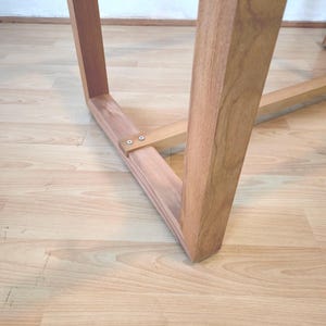 K&ouml;nnte beinhalten: Nahaufnahme eines Holz-Tischbeins und der Tragstruktur. Das Bein ist hellbraun und abgewinkelt, mit rechteckiger Form. Die Halterung ist mit kleinen Schrauben befestigt. Der Tisch steht auf einem hellen Holzboden.