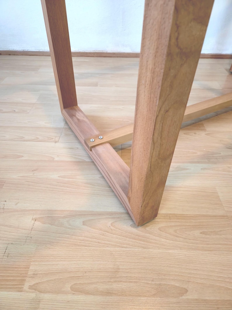 K&ouml;nnte beinhalten: Nahaufnahme eines Holz-Tischbeins und der Tragstruktur. Das Bein ist hellbraun und abgewinkelt, mit rechteckiger Form. Die Halterung ist mit kleinen Schrauben befestigt. Der Tisch steht auf einem hellen Holzboden.