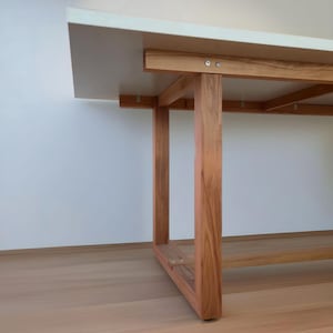 K&ouml;nnte beinhalten: Ein moderner rechteckiger Tisch mit wei&szlig;er Tischplatte und Beinen aus Naturholz. Die Tischbeine haben eine warme braune Farbe, und die Tischplatte scheint eine glatte, solide Oberfl&auml;che zu sein. Der Tisch ist ein modernes Design.