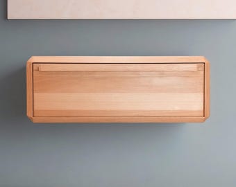 May include: A light-colored wooden floating drawer with a rectangular shape. The drawer is mounted on a blue-gray wall. Above the drawer is a white rectangular shape.