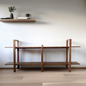 May include: A wooden shelving unit with two shelves and a floating shelf above. The shelves are made of dark wood and are empty. A black vase with greenery and a small potted plant sit on the shelves. The room has light wood flooring and white walls.