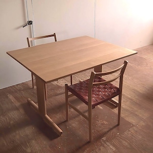 Puede incluir: Una mesa de madera con un acabado en madera clara y dos sillas. Una silla tiene un asiento de cuero tejido de color marrón rojizo.