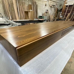 Peut inclure: Un grand meuble rectangulaire en bois avec une finition marron riche. Le meuble pr&eacute;sente un design &eacute;pur&eacute; et moderne avec une surface lisse et polie et une section encastr&eacute;e avec des rainures horizontales. Pos&eacute; sur une surface blanche dans un atelier.