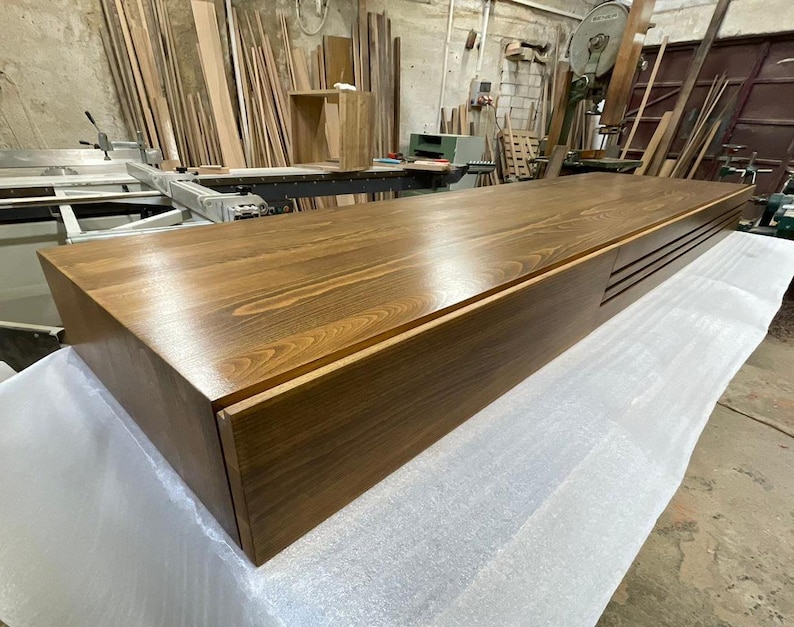 Peut inclure: Un grand meuble rectangulaire en bois avec une finition marron riche. Le meuble pr&eacute;sente un design &eacute;pur&eacute; et moderne avec une surface lisse et polie et une section encastr&eacute;e avec des rainures horizontales. Pos&eacute; sur une surface blanche dans un atelier.