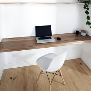 May include: A white desk chair with black legs sits in front of a floating wooden desk with a laptop computer on top. The desk is mounted to a white wall with a wooden shelf above it.
