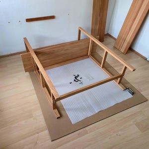 May include: Wooden furniture frame in the process of assembly, with various wooden components and hardware visible. The frame is light brown, and sits on a cardboard sheet. Additional wooden planks are leaning against a wall in the background.