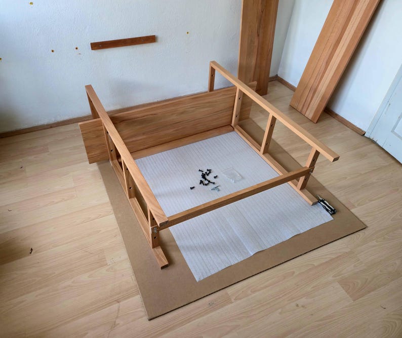 May include: Wooden furniture frame in the process of assembly, with various wooden components and hardware visible. The frame is light brown, and sits on a cardboard sheet. Additional wooden planks are leaning against a wall in the background.