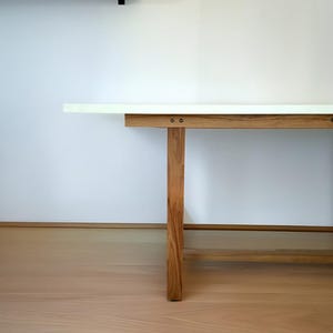 K&ouml;nnte beinhalten: Ein moderner Holztisch mit wei&szlig;er Tischplatte. Der Tisch hat vier stabile Holzbeine und eine hellbraune Oberfl&auml;che. Der Tisch steht vor einer wei&szlig;en Wand und einem hellbraunen Holzboden.