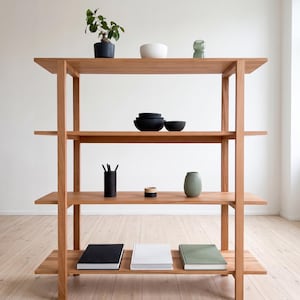 May include: A light-colored wooden bookshelf with four shelves. The shelves display various items, including a potted plant, bowls, books, and decorative objects. The bookshelf is set against a white wall and light-colored wooden floor.