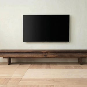May include: A modern living room featuring a large, dark screen television mounted above a low, wooden media console. The console is approximately 72 inches long. A potted plant and a gray armchair are also visible.