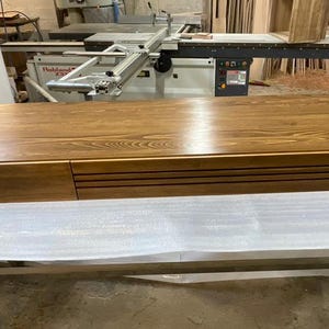 Peut inclure: Un grand meuble rectangulaire en bois avec une finition en grain de bois naturel. Le meuble a un tiroir sur le c&ocirc;t&eacute; gauche et une s&eacute;rie de rainures horizontales sur le c&ocirc;t&eacute; droit. Il repose sur un cadre m&eacute;tallique recouvert d'un mat&eacute;riau protecteur blanc.