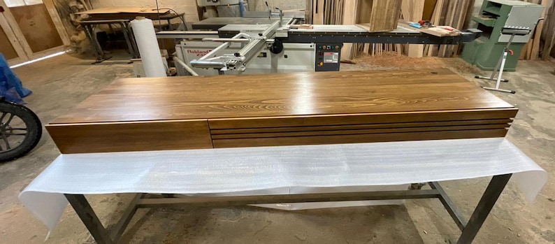 Peut inclure: Un grand meuble rectangulaire en bois avec une finition en grain de bois naturel. Le meuble a un tiroir sur le c&ocirc;t&eacute; gauche et une s&eacute;rie de rainures horizontales sur le c&ocirc;t&eacute; droit. Il repose sur un cadre m&eacute;tallique recouvert d'un mat&eacute;riau protecteur blanc.