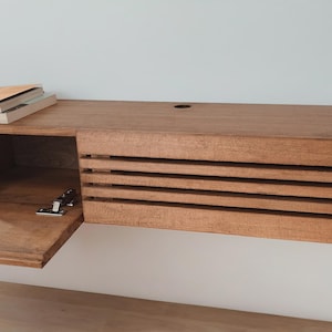 May include: A wooden wall-mounted shelf with a hinged door that opens to reveal an empty storage space. The shelf has a slatted front and is made of a light brown wood.