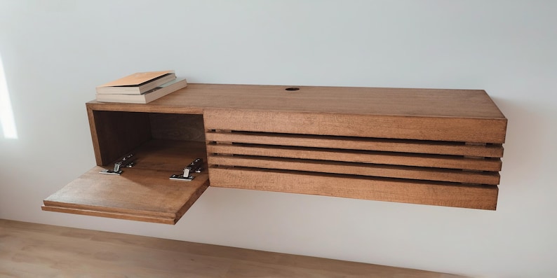 May include: A wooden wall-mounted shelf with a hinged door that opens to reveal an empty storage space. The shelf has a slatted front and is made of a light brown wood.