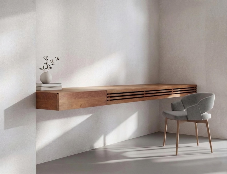 Peut inclure: Un bureau flottant moderne en bois clair, avec un tiroir et des lattes, associ&eacute; &agrave; une chaise grise rembourr&eacute;e. Le bureau est fix&eacute; &agrave; un mur blanc. Un petit vase avec de la verdure et des livres sont sur le bureau.
