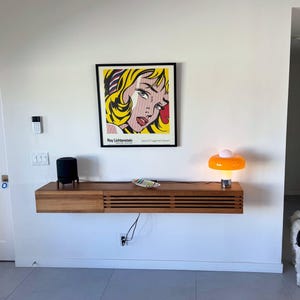 May include: A floating shelf with a black speaker, a small plate, and an orange and white lamp. A framed print of a comic book style woman's face by Roy Lichtenstein hangs above the shelf. A white dog is standing in the doorway.